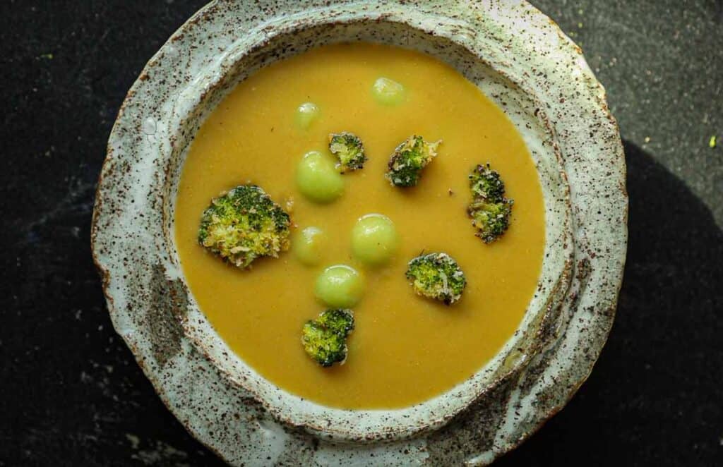 A speckled ceramic bowl filled with creamy broccoli soup garnished with broccoli florets and small green spheres. The bowl is set against a dark background.